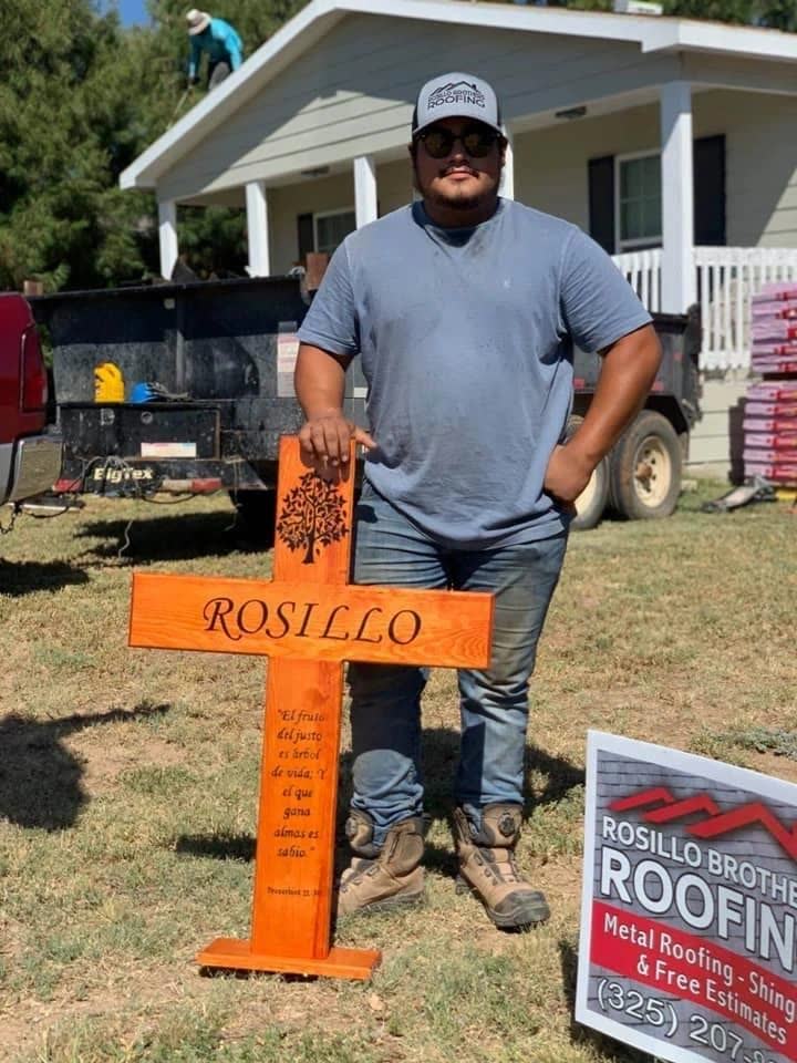 A man on a Rosillo job holding an orange wooden cross with ROSILLO carved on it, next to the company's yard sign